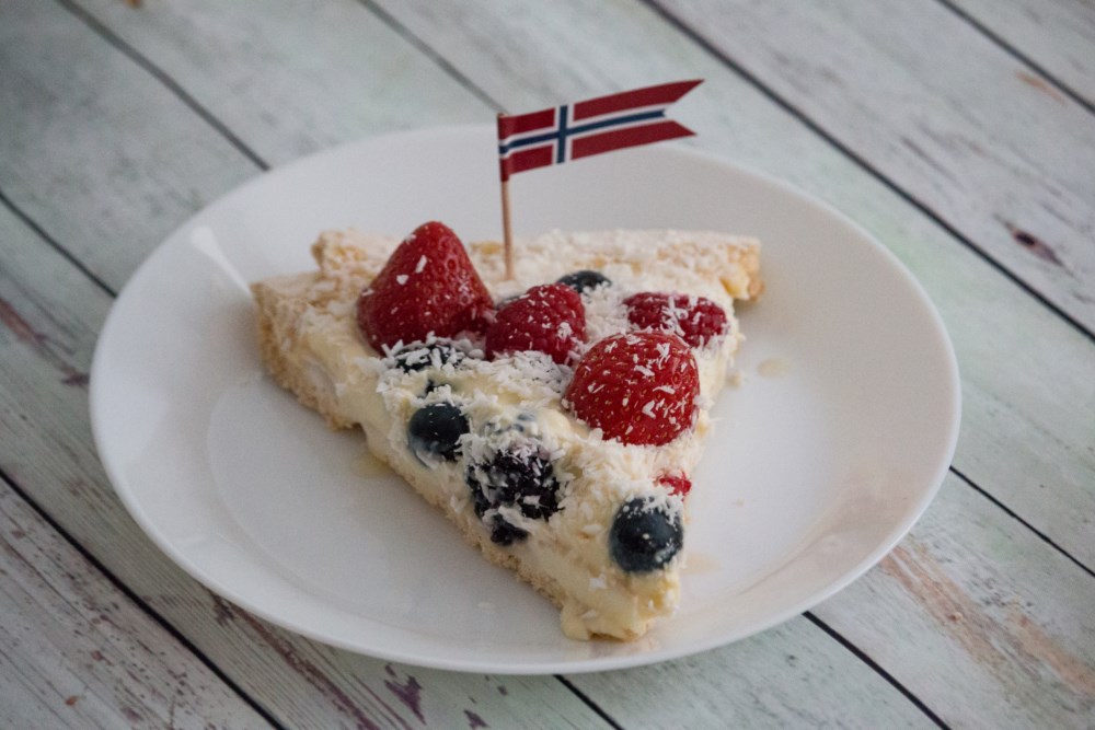 Et kakestykke pyntet med friske bær og et lite norsk flagg på toppen, klart til feiring på 17. mai.